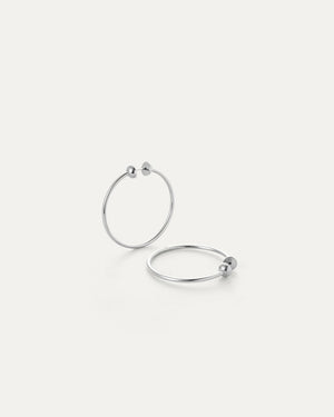 Two JENNY BIRD SMALL ICON HOOP EARRINGS crafted from stainless steel lie on a light gray surface. Featuring a minimalist design with a spherical accent, the clean backdrop highlights their sleek elegance and timeless appeal.