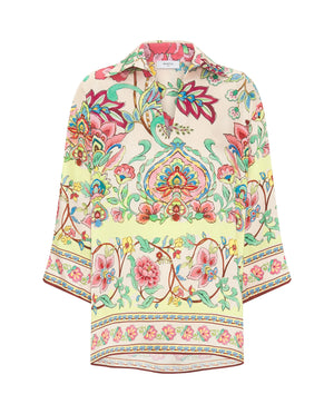 A person with long blonde hair stands against a plain background, wearing the MIRTO 1956 - FLUID BLOUSE MULTICOLOR FLORAL by MIRTO 1956. The uplifting blouse features a vibrant, floral pattern in pink, green, yellow, and blue. The 3/4 sleeves and V neckline enhance the intricate designs, creating a bright and stylish look.