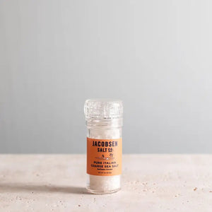 A clear salt grinder containing coarse Italian sea salt from Trapani is displayed against a plain white background. The label on the JACOBSEN SALT - GRINDER COARSE ITALIAN SEA SALT provides the ingredients (sea salt), manufacturer details, and a URL. The JACOBSEN SALT CO. grinder features a transparent top for dispensing the salt.