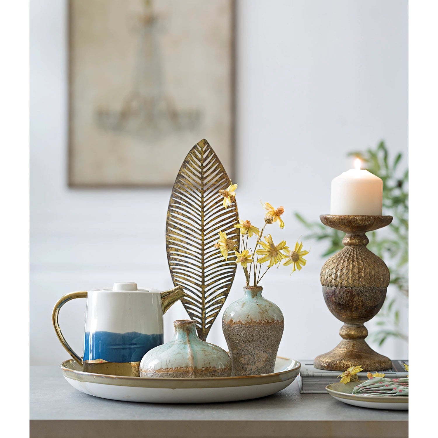 On a round tray, a ceramic tea set with blue accents, two CREATIVE COOP DISTRESSED TERRACOTTA VASES - SMALL holding yellow flowers, a gold leaf sculpture, and a lit candle on a textured holder are arranged decoratively, with blurred wall art in the background.