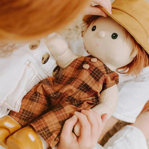 A Dinkum Doll from Olli Ella USA's Travel Togs collection, featuring dark hair and dressed in a denim jacket, mustard pants, and black shoes, stands on a wooden surface. Beside the doll is a teal See-Ya Suitcase with white trim and hardware. In the background, another larger teal suitcase is partially visible.