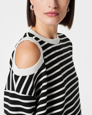 A person with short hair poses against a white background, wearing the SPANX - AIRESSENTIALS COLD SHOULDER TOP in black and white stripes crafted from luxuriously soft material, paired with light-colored pants. Their left hand is placed on their head while their right arm rests at their side.