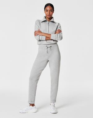 A person stands against a white background, smiling and looking down with one hand touching their hair. They are wearing a matching light beige SPANX AIRESSENTIALS TAPERED PANT and sweatshirt set made from ultra drapey, lightweight spacer fabric, paired with white sneakers.