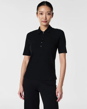 A woman in the SPANX - STRETCH RIB POLO TOP, a brown, ribbed, short-sleeve shirt with a button-down front, stands with her arms relaxed. The plain white background accentuates the top's stretchy ribbed fabric and body-hugging fit.