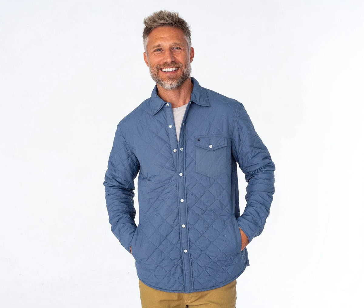 A man with a gray beard wears the CRIQUET - QUILTED SHACKET, a light blue piece with a pearl snap closure, along with a cream cap. He stands outdoors among pine trees and greenery in a forested area.