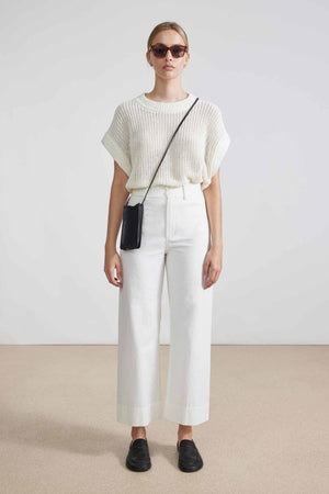 A person with hair in a bun is facing away, wearing sunglasses, a cream knit top, and the APIECE APART - CLASSIC MERIDA PANT that creates a sleek silhouette. Black shoes and a thin black crossbody strap complete the look against a light, minimalist setting.