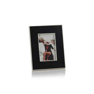 Two framed photographs are displayed on a textured surface. One MDF frame, slightly larger, holds a photo of a person resting on a surface. The smaller frame, from Zodax's BLACK BONE INLAY WITH WHITE BORDER 4X6 collection, contains an image of a person sitting and wearing a hat. Both frames have dark borders with light interiors.