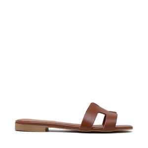 The image depicts the MICHELE LOPRIORE - H SLIDE, a single brown leather slide sandal with an open-toe design. The upper features two wide straps with geometric cutouts. The thin sole appears to be made of light brown wood-like material. Made in Italy by MICHELE LOPRIORE, the sandal is displayed against a plain white background.