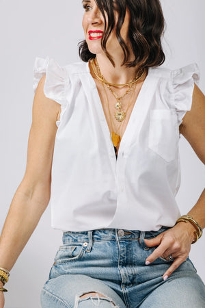 A woman with shoulder-length dark hair is seen sitting in a relaxed pose, wearing the effortlessly chic KMJ - Olivia Sleeveless with Ruffle—a white blouse featuring ruffles on the shoulders—paired with layered gold necklaces and gold bracelets. She completes her look with blue jeans that have a slight rip on the knee.