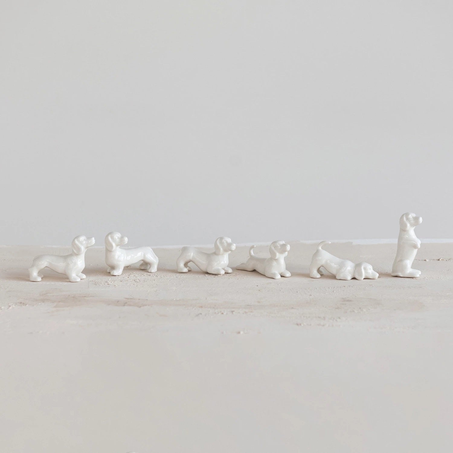 The STONEWARE DOGS - SET OF 6 by CREATIVE COOP features six small white ceramic dachshunds in various poses like crouching, standing, and lying down, arranged on a light surface against an off-white background.