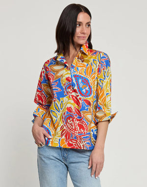 A woman with shoulder-length dark hair is shown from behind, wearing the HINSON WU Aileen 3/4 Sleeve Paisley Print Top, which features a colorful blend of red, yellow, blue, and orange shades. She has paired it with light blue denim jeans against a plain white background. This best-selling top from HINSON WU exudes style and vibrancy.