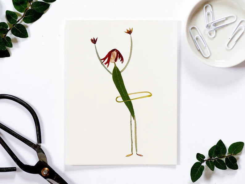 The "CARD HULA HOOP" by PETAL PEOPLE PRESS is a minimalist botanical card featuring a stick figure ballerina dressed in green with red hair, surrounded by leaves. To the left are black scissors, and to the right is a small white dish with paperclips, adding an element of whimsy to this distinctive piece.