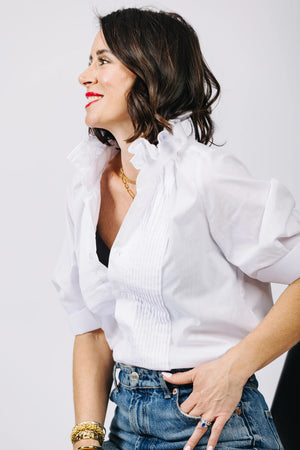 A woman with blond hair, wearing KMJ's SOUTHERN LOVER TUX adorned with pleated detailing and a turquoise necklace, is adjusting the waistband of her Demi-Boyfriend blue jeans. The background is plain white.