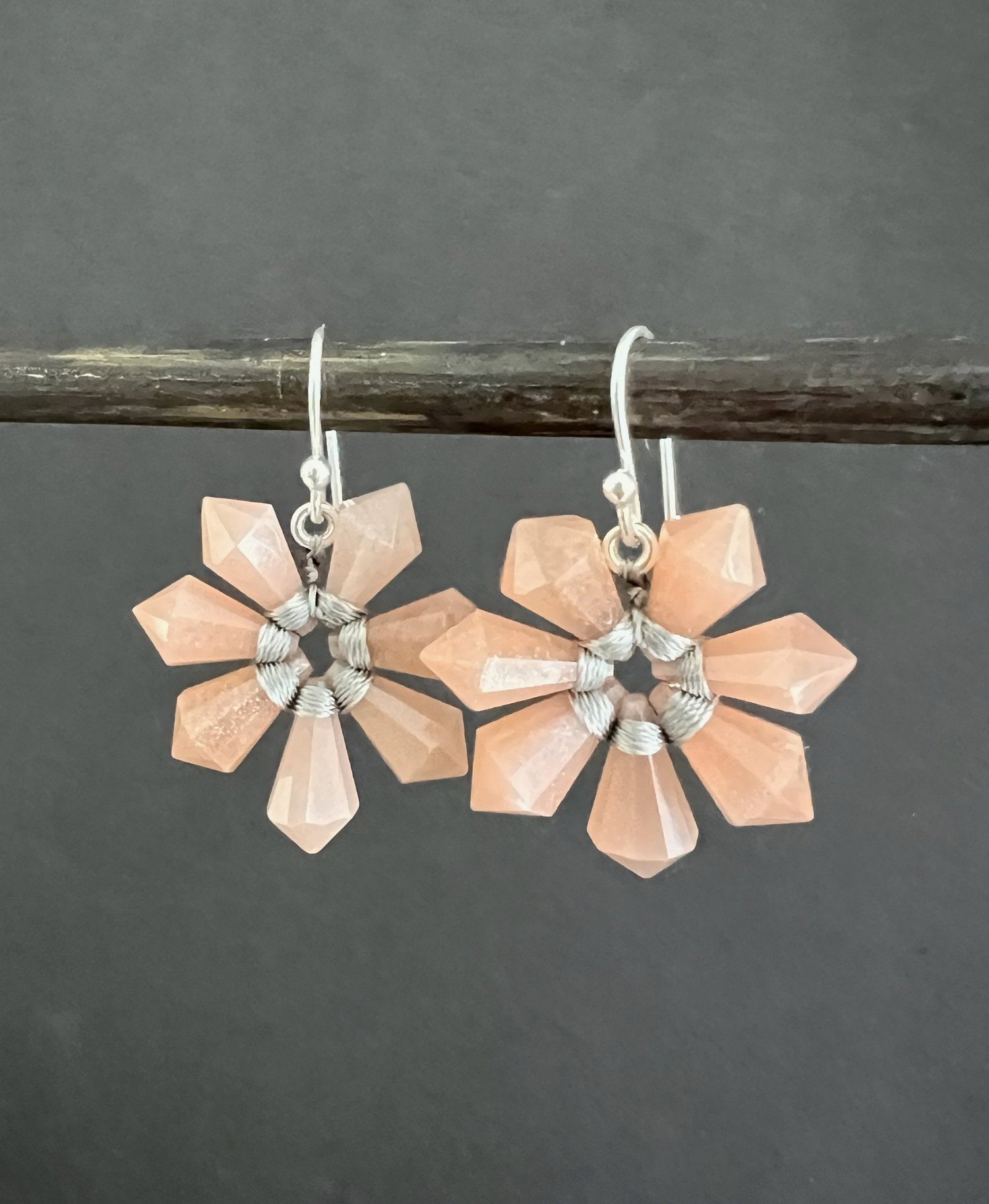 Two ANN LIGHTFOOT SEVEN POINT FACETED PINK MOONSTONE FLOWER EARRINGS hang from a dark rod, featuring faceted petals and a sterling silver weave, in a symmetrical design against a plain dark gray backdrop.