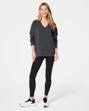 A person with curly hair is standing and looking to the side, wearing a cozy SPANX - AIRESSENTIALS BRUSHED V-NECK TUNIC paired with brown pants. The breathable top drapes elegantly, capturing a sense of laid-back comfort against the plain, light-colored background.