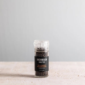 A JACOBSEN SALT CO. grinder, filled with Tellicherry peppercorns, sits gracefully on a light-colored surface against a plain, light gray background. The clear glass bottle bears a striking label in black with white and orange text.