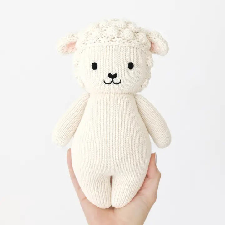 Introducing the CUDDLE + KIND - HANDMADE BABY LAMB by CUDDLE &amp; KIND. This delightful knitted stuffed toy is crafted from 100% cotton yarn and features a white, wool-like texture. It has adorable black eyes, a smiling mouth, and small round ears. The toy’s body boasts a simple design with a smooth and plush finish. Perfect as one of your fair trade baby gifts!