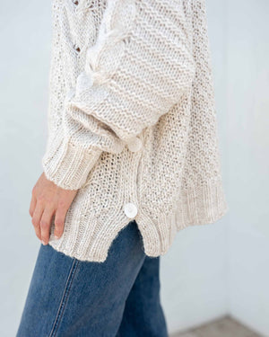 The image displays a detailed view of a person dressed in the MERSEA - TOPANGA CABLE SWEATER by MER SEA, showcasing its chunky knit cream-colored texture with large buttons on the sides, paired with blue jeans. The emphasis is on the sweater's intricate cable design and fine details, with the person's hand partially visible at their side.