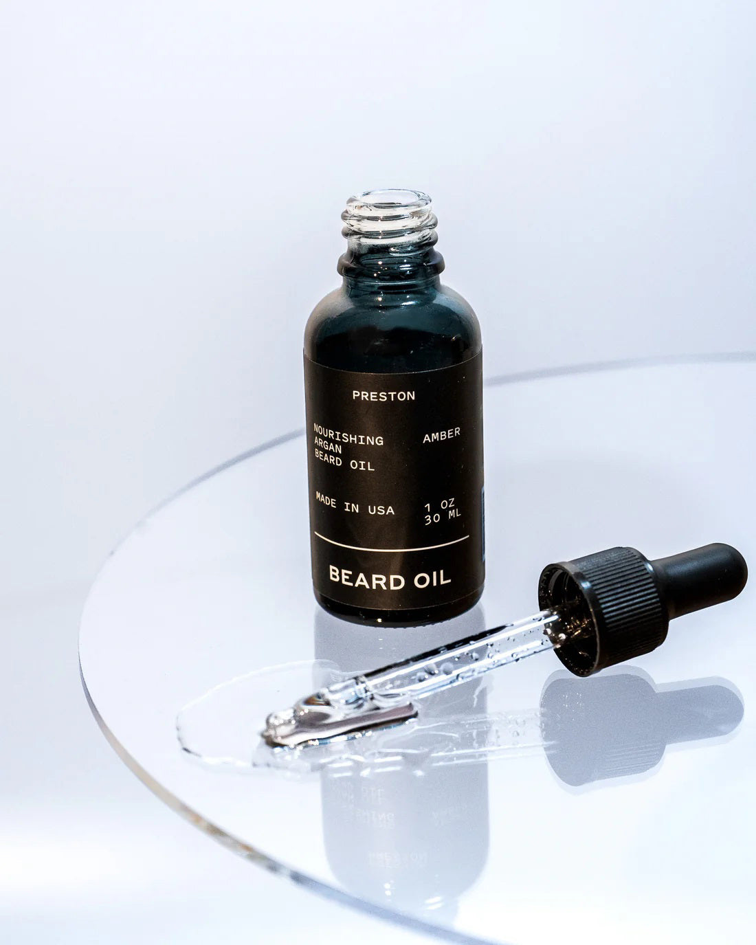 A black glass bottle labeled &quot;PRESTON - BEARD OIL&quot; is placed on a reflective surface. A dropper with a black rubber top and glass pipette lies beside it, with some argan oil visible around the pipette.