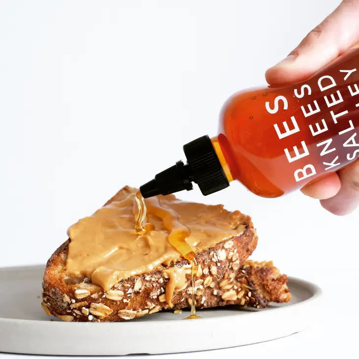 A red bottle of BUSHWICK KITCHEN - 'BEES KNEES' SALTED HONEY is shown. The label reads "BORN IN BROOKLYN, NY" and "NET WT 12.5 OZ (354g)." This gluten-free treat features wildflower honeys perfected with a hint of salt and comes with a black pointed cap.