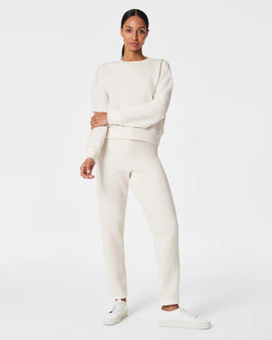 A woman stands confidently against a plain white background, wearing the SPANX Aireessentials Crew Neck Pullover and matching sweatpants made from luxurious comfort spacer fabric. She has her hair pulled back and her left hand is raised slightly. She smiles gently with her lips closed.