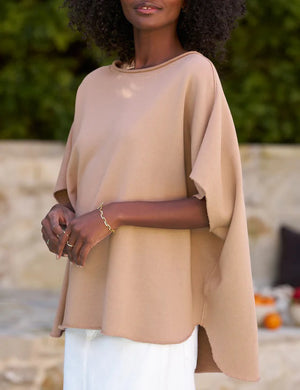 A woman with voluminous curly hair walks outdoors. She is wearing a loose-fitting FRANK AND EILEEN - OLIVE ORIGINAL CAPELET and white trousers, reminiscent of an original capelet. Behind her are lush green trees and a stone wall with a cushioned bench. She has a calm and confident expression.