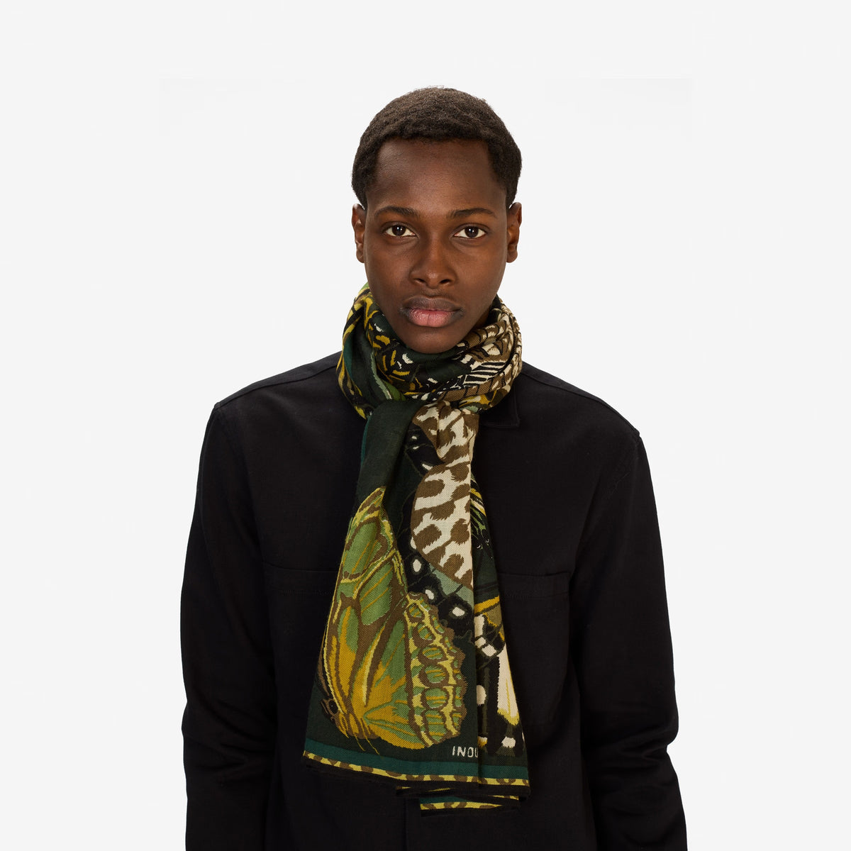 A woman in a light gray sweater and black pants stands against a plain white background, wearing the INOUI EDITIONS - PAPILLON (BUTTERFLIES) wool scarf in emerald from INOUI, featuring black and yellow butterfly designs.