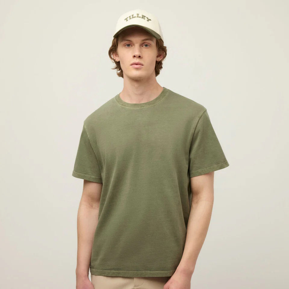 A young man with light skin and short brown hair wears the TILLEY Mid Weight Sunwashed T-Shirt in Thyme Green and a cream &quot;TILLEY&quot; baseball cap against a light, neutral background.