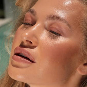 A close-up of a person with curly dark hair, a serious expression, and warm makeup featuring the universally flattering, weightless JASON WU - HOT FLUFF LIP/EYE/CHEEK by JASON WU BEAUTY on their face. They are looking directly at the camera with a neutral background.