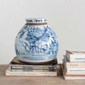 A round, distressed blue-and-white ceramic SONEWARE GINGER JAR by CREATIVE COOP with floral and nature designs sits on a wooden table. The jar is placed on top of a stack of books, including titles like "Toulouse-Lautrec Paintings" and "1941 Drawings Year." The background is plain and off-white.