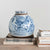 A round, distressed blue-and-white ceramic SONEWARE GINGER JAR by CREATIVE COOP with floral and nature designs sits on a wooden table. The jar is placed on top of a stack of books, including titles like "Toulouse-Lautrec Paintings" and "1941 Drawings Year." The background is plain and off-white.