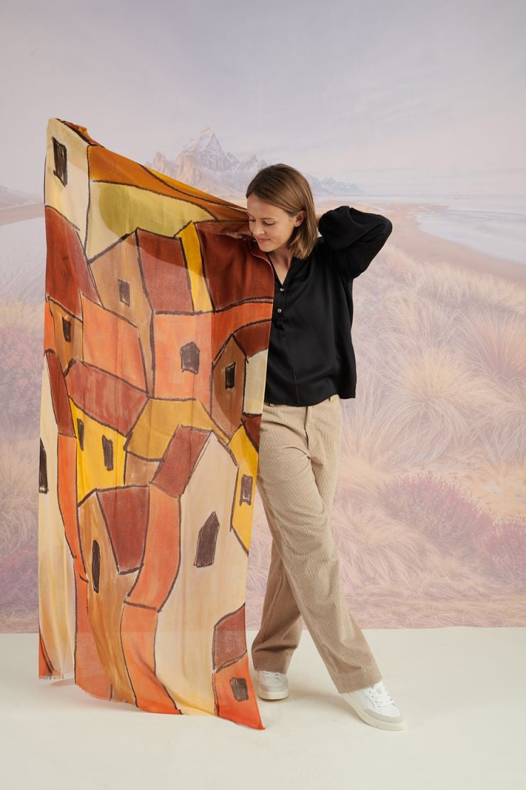 A woman in a black long-sleeve top and beige pants stands before a scenic backdrop, her look elevated by the STORIATIPIC ISCHIA Modal and Wool Scarf (100x200 cm) in vibrant orange and brown, draped elegantly around her neck.