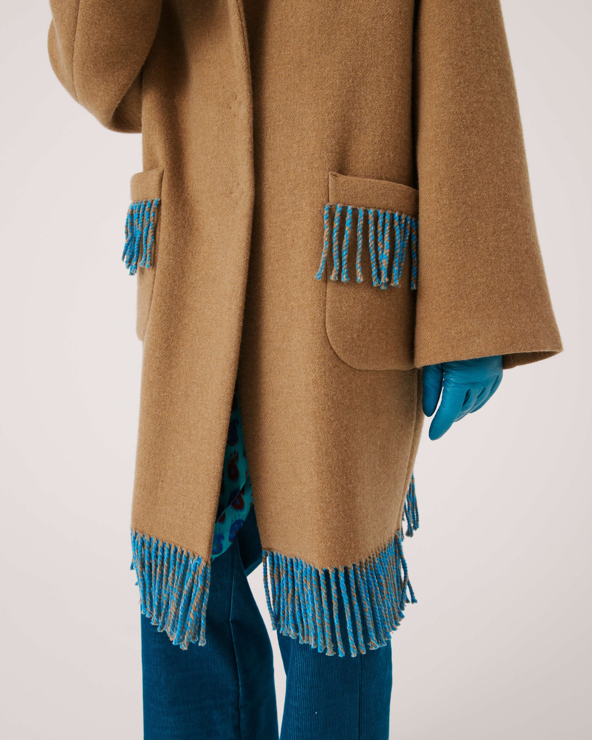 A person is wearing the SFIZIO - TWO TONED COAT WITH FRINGE by SFIZIO, featuring blue and brown fringe patch pockets and hem. They are also donning blue gloves and blue pants. The image is cropped to show from the shoulders down against a plain, off-white background.