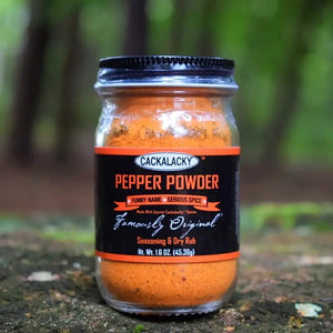 A glass jar of CACKALACKY - PEPPER POWDER RUB, made with fiery chile peppers and secret spices, is shown against a white background. The jar has a black lid and a black label with orange and white text stating "Cackalacky Pepper Powder," "Funny Name - Serious Spice," and "Famously Original." The jar contains 2.3 oz (65.2g).