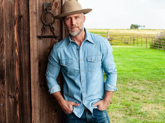 A man with a gray beard wears the CRIQUET WESTERN DENIM SHIRT WITH A PEARL SNAP IN LIGHT WASH by CRIQUET as he stands outdoors near a metal fence, with green grass and an open field in the background.