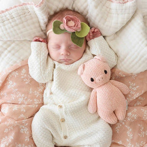 A pink knitted baby piglet, crafted from 100% cotton yarn, is shown from the back. It has small round ears, arms, a tail, and a tag attached to its side. The piglet boasts a simple, cuddly design and radiates the charm of hand-knit baby animals while supporting fair trade income. This delightful creation is known as the CUDDLE + KIND - HANDMADE BABY PIGLET by CUDDLE & KIND.