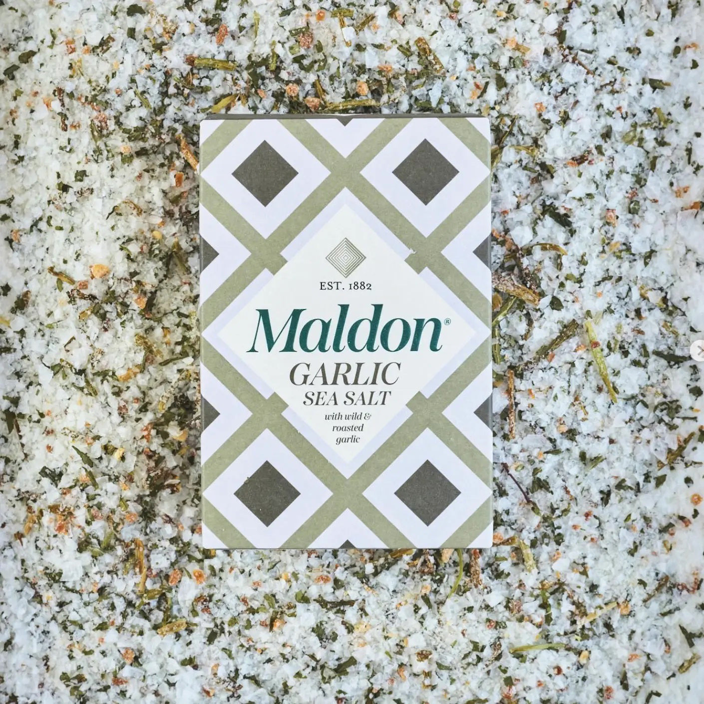 A box of GOURMET FOOD SOLUTIONS' MALDON - GARLIC SEA SALT is placed on a bed of coarse salt with herbs and garlic flakes scattered around. The box, in a green and white geometric design, reads "MALDON - GARLIC SEA SALT" and "with white & roasted garlic," established 1882—proudly showcasing its English sea salt heritage.