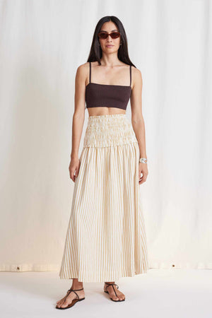 A woman with long hair is wearing sunglasses, a dark crop top, and the APIECE APART - ORA SMOCK MAXI SKIRT made of organic cotton. She is standing against a light-colored background and wearing sandals.