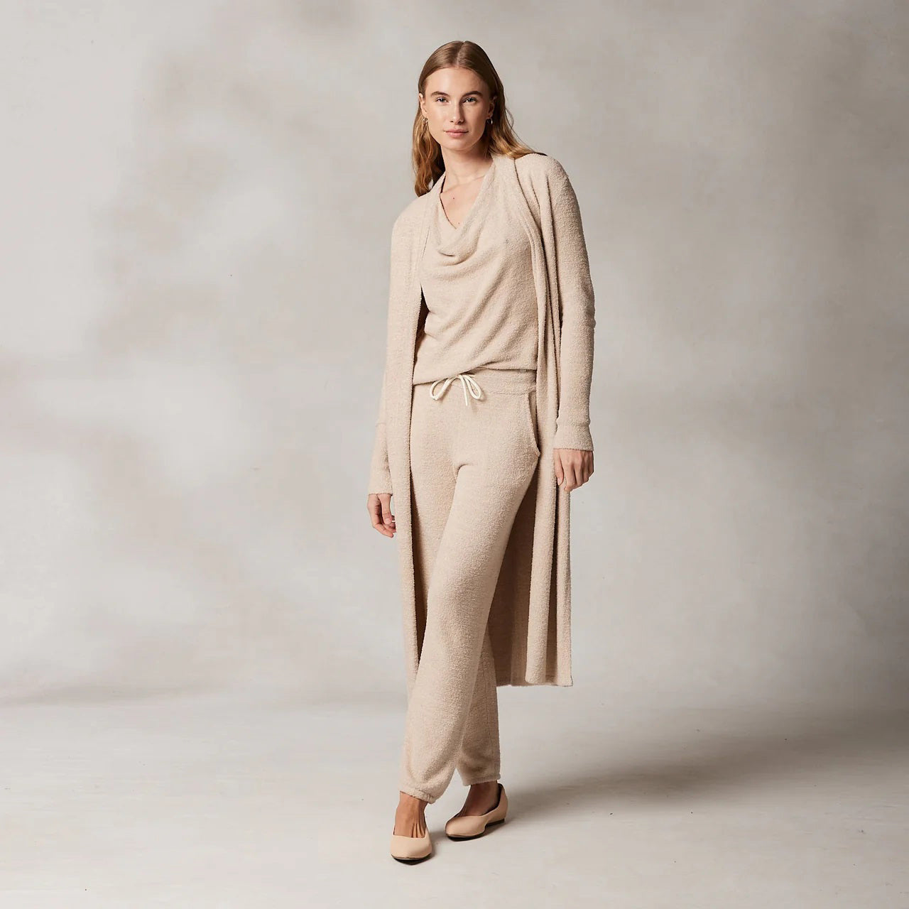 A woman with long hair wearing the KASHWERE - LONG CARDIGAN, part of a cozy beige lounge set from KASHWERE, crafted from Kash-ease yarn. The ensemble consists of a long-sleeved top, drawstring pants, and the matching long cardigan. She stands against a light grey background, looking confidently at the camera in her laid-back luxury outfit. Beige flat shoes complete the look.