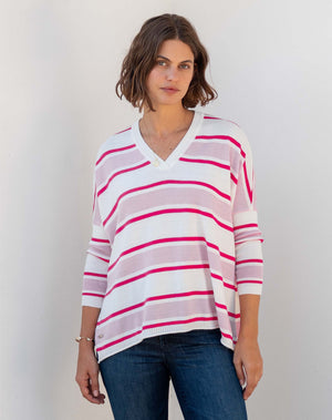 A person with short curly hair is standing against a plain white background. They are wearing a loose-fitting MER SEA - CATALINA V-NECK SWEATER with green diagonal stripes and blue jeans. This versatile year-round closet hero, featuring MERSEA's signature side slits, complements their look perfectly. The person gazes directly at the camera with a neutral expression.