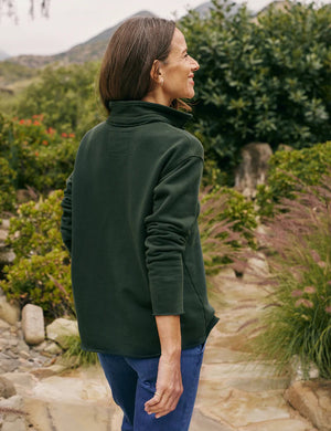 A person with long brown hair wearing a dark green FRANK & EILEEN - Patrick Popover Henley in Triple Fleece Evergreen stands outdoors. They are adjusting the collar of their top with both hands. The background is lush with greenery and soft-focus plants.
