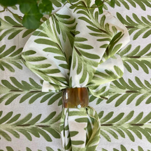 The CHRISTINA DICKSON HOME FERN COTTON NAPKIN, featuring a green leaf block print on 100% cotton and held by a brown ring, complements the matching tablecloth beneath with its seamless leaf motif design. A small green plant is visible in the top left corner.