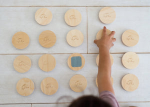 A young boy in a green shirt plays intently with the LITTLE MATCHABLES GAME SET by LILY & RIVER on a white table in a Montessori playroom. The room is adorned with various toys and baskets, and the boy appears focused on enhancing his fine motor skills by arranging and stacking the round wooden discs as part of this engaging memory game.