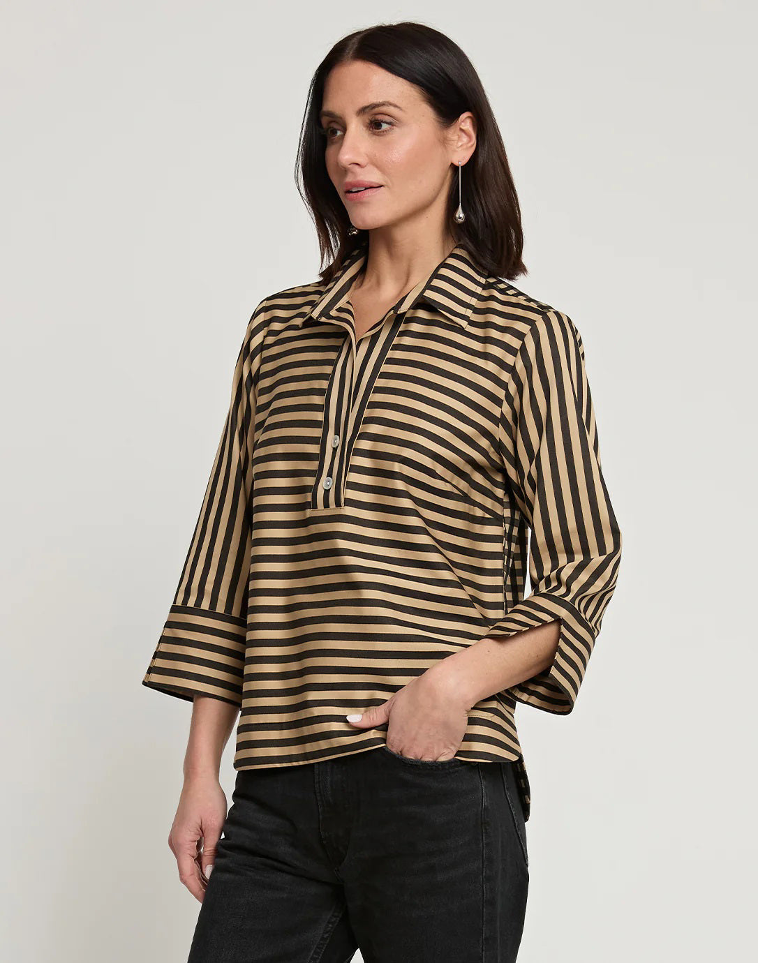 A woman with shoulder-length dark hair wears the HINSON WU - AILEEN 3/4 SLEEVE STRIPE GINGHAM COMBO SHIRT, a black and beige striped blouse featuring a small collar and a partial zip at the front. She pairs it with black pants and stands against a plain white background, one hand in her pocket.