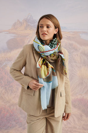 A woman with long blonde hair, in a light blue shirt and dark pants, smiles in front of a scenic backdrop, draping the elegant, hand-embroidered STORIATIPIC - ASTER WOOL SCARF around her shoulders.