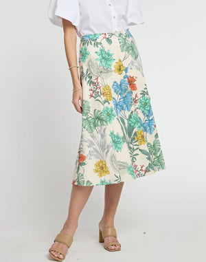 A person is wearing a white button-up shirt with short, puffy sleeves, and a high-waisted GLORIA SKIRT by HINSON WU featuring a vibrant Rainforest Print in various colors. They are also wearing tan, open-toe sandals with a low heel. The background is plain white.