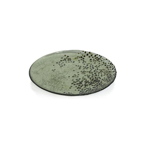 A round, light green DOTTED GLASS PLATE 8.25" from ZODAX features an uneven surface with a dark speckled pattern radiating from one edge, giving it a slightly rustic, handmade appearance. Photographed against a plain white background, its dimensions emphasize the artisanal charm of this distinctive piece.