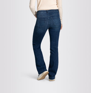 A person wearing a long-sleeve, light-colored ribbed top and MAC's DREAM BOOT AUTHENTIC DENIM boot cut jeans in dark blue with a slight flare at the bottom. They are also wearing white sneakers and have one hand in their pocket, standing against a plain, light gray background.