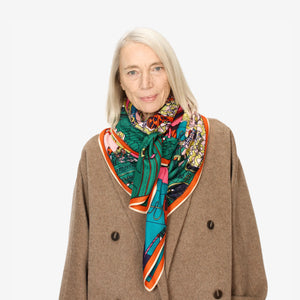 A person with long light gray hair is wearing a brown coat and the INOUI EDITIONS - IMPRIMERIE SQUARE SCARF by INOUI around their neck. The scarf showcases vibrant hues of green, blue, red, and orange. The person is standing against a plain white background.