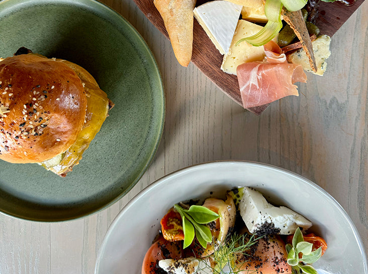 A top-down view of a table with a sandwich bun on a green plate, a charcuterie board with cheese, cured meats, and crackers, and a salad with tomatoes, herbs, and cheese on a white plate.